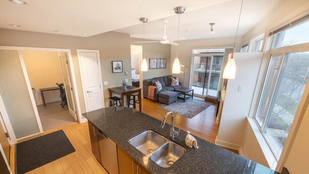 A modern kitchen with granite countertops, stainless steel sinks, and a dining area with a gray sofa and coffee table. - Mt. Tolmie View-Spacious, Bright NE Facing Two Bedroom Condo with Den by Victoria Prime! (Victoria)