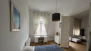 Ein Schlafzimmer mit Bett, Schreibtisch, Stuhl, einem Fenster mit Vorhängen, einer Pendelleuchte und einem Fernseher.