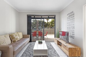 A modern living room with a sofa, coffee table, TV, and a sliding glass door leading to an outdoor patio with red chairs and a table. - Waterstone East C103 (Cape Town)