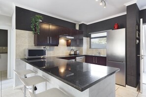 A modern kitchen with dark wooden cabinets, a stainless steel refrigerator, and a large island with bar stools. - Waterstone East C103 (Cape Town)