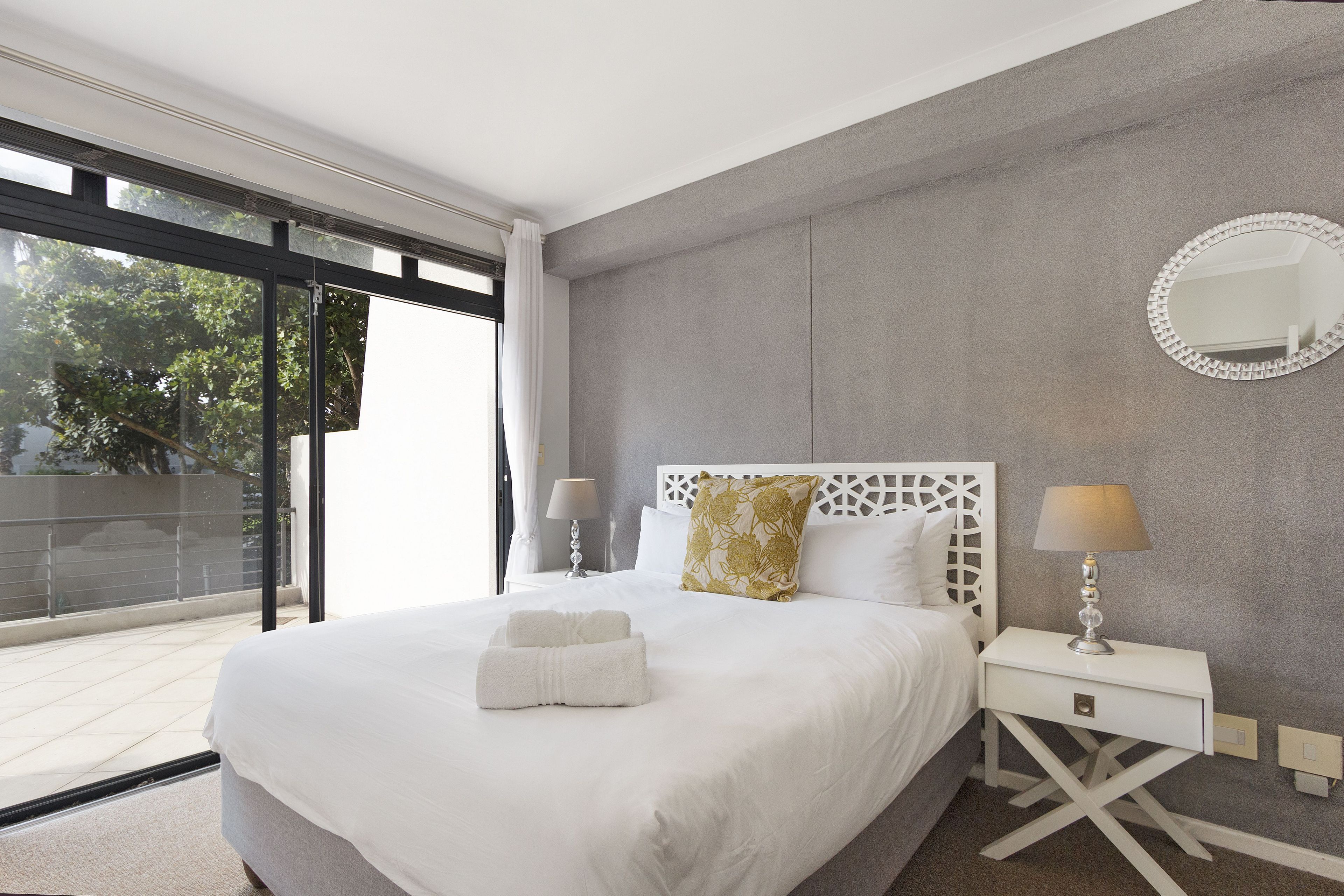 Une chambre moderne avec un grand lit, deux lampes de chevet, un miroir rond et une vue dégagée sur l’extérieur grâce à une grande fenêtre.