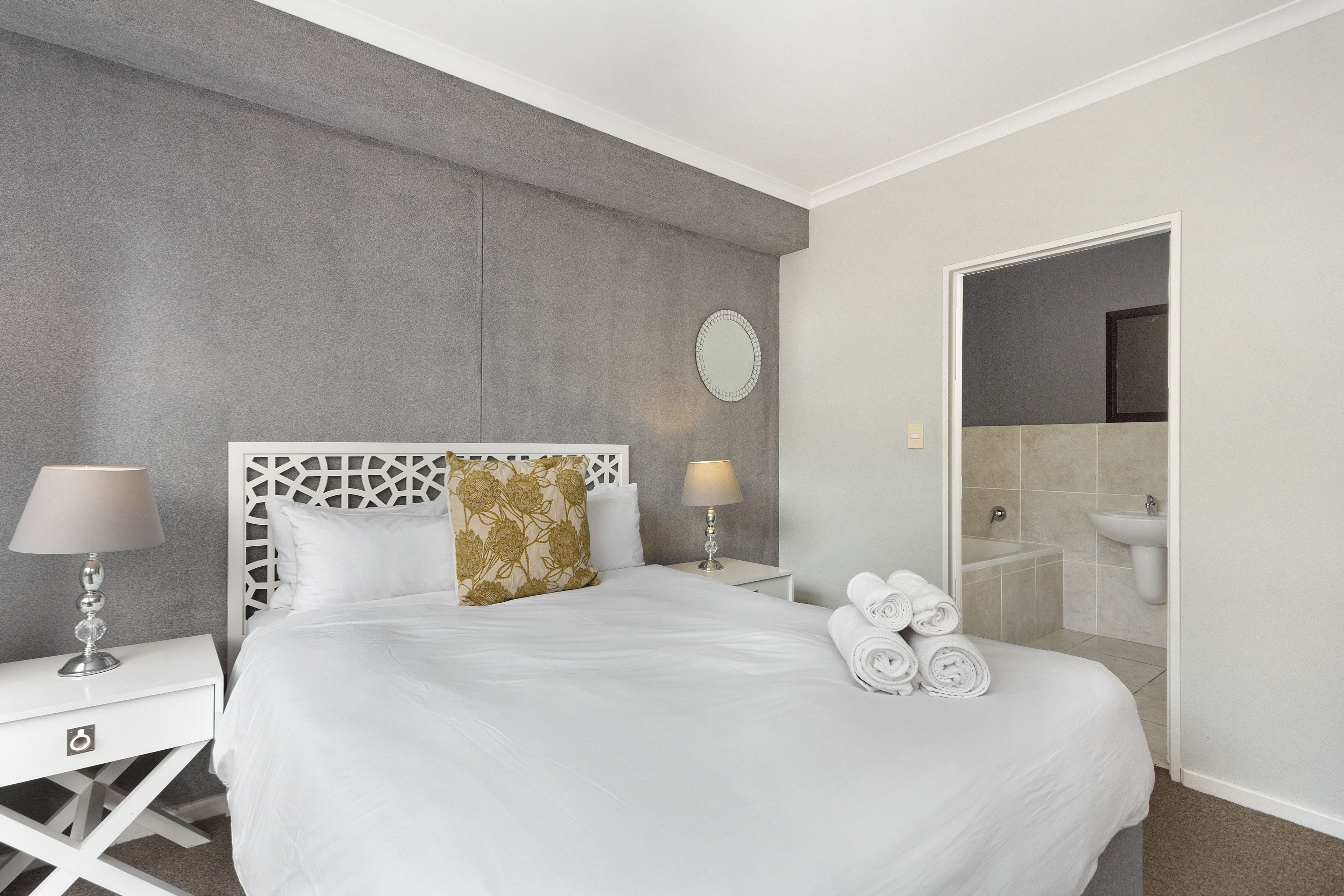 Une chambre d’hôtel avec un lit, deux tables de chevet, un miroir et une salle de bain visible à travers une porte ouverte.