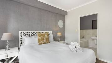 Une chambre d’hôtel avec un lit, deux tables de chevet, un miroir et une salle de bain visible à travers une porte ouverte.