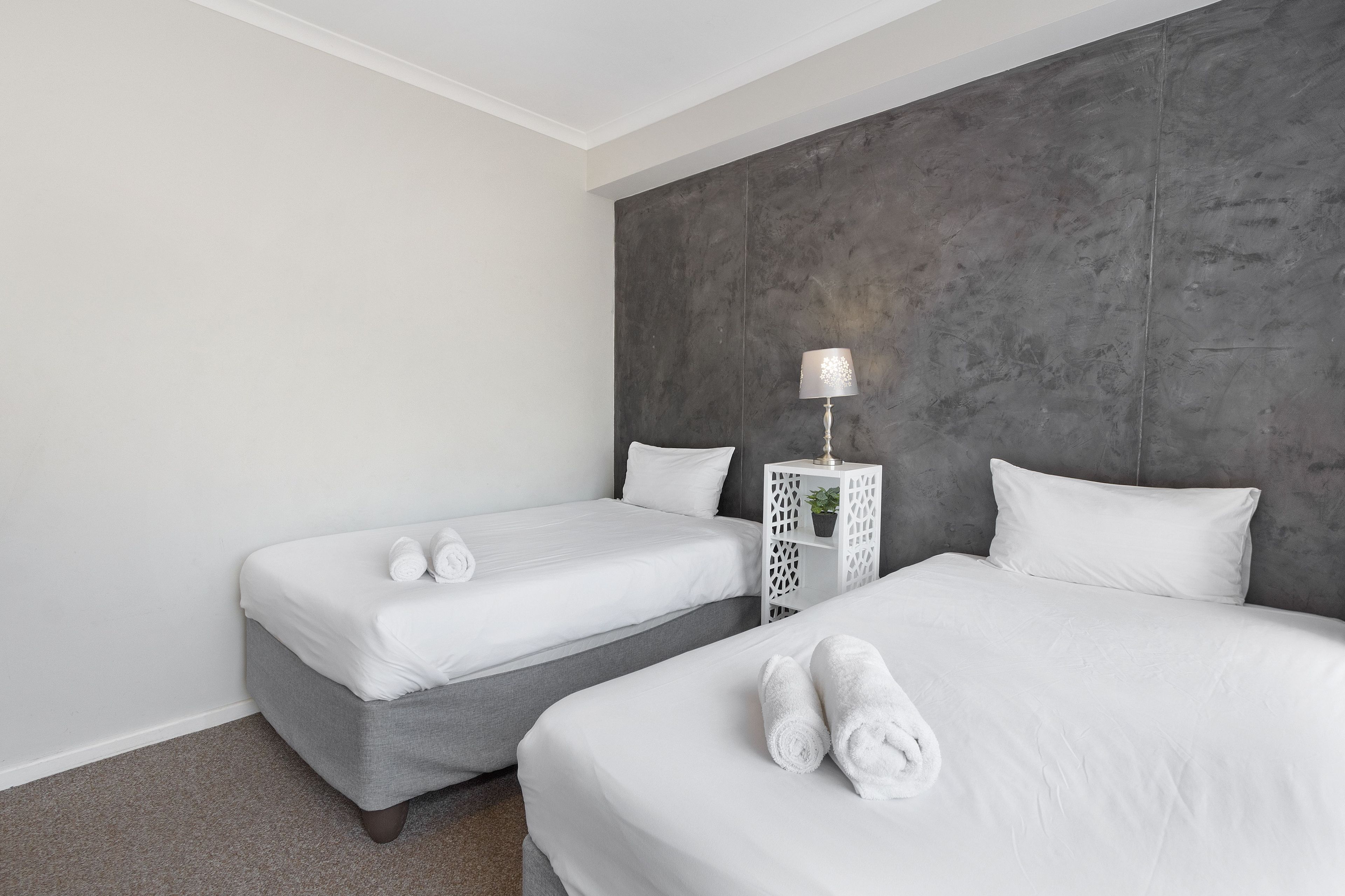 A hotel room with two beds, a grey accent wall, and a white nightstand with a lamp.