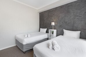 A hotel room with two beds, a grey accent wall, and a white nightstand with a lamp. - Waterstone East C103 (Cape Town)