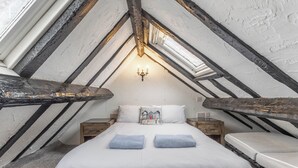 Ein Schlafzimmer mit schrägverdeckter Decke, freiliegenden Holzbalken, einem Bett mit zwei Kissen, einem Nachttisch und einer Wandleuchte.