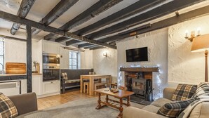 Ein gemütliches Wohnzimmer mit Kamin, Holzbalken, einem Fernseher und einer Küchenzeile mit modernen Geräten.
