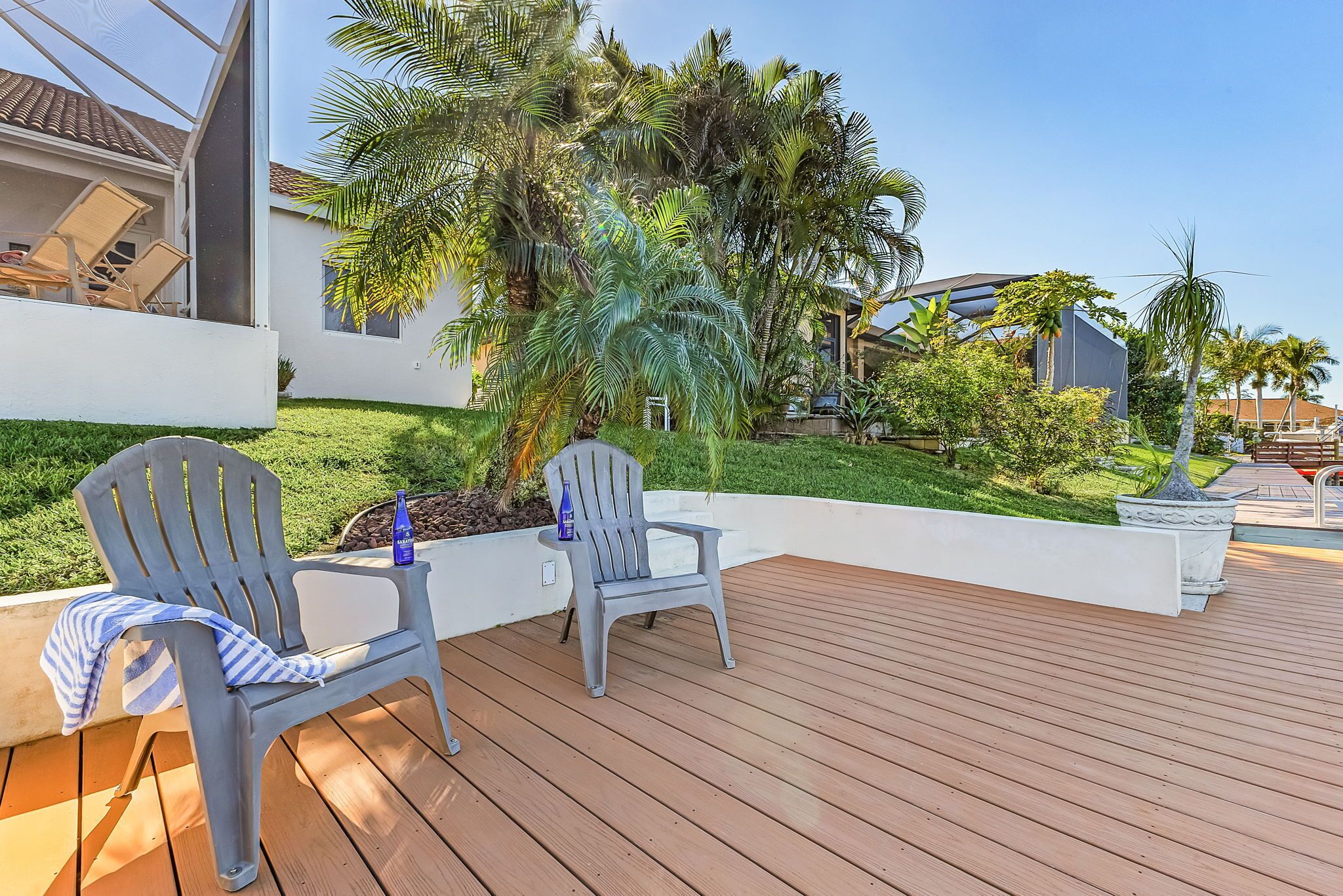 Une terrasse en bois avec deux chaises grises et une bouteille, entourée d’une végétation luxuriante et de palmiers.