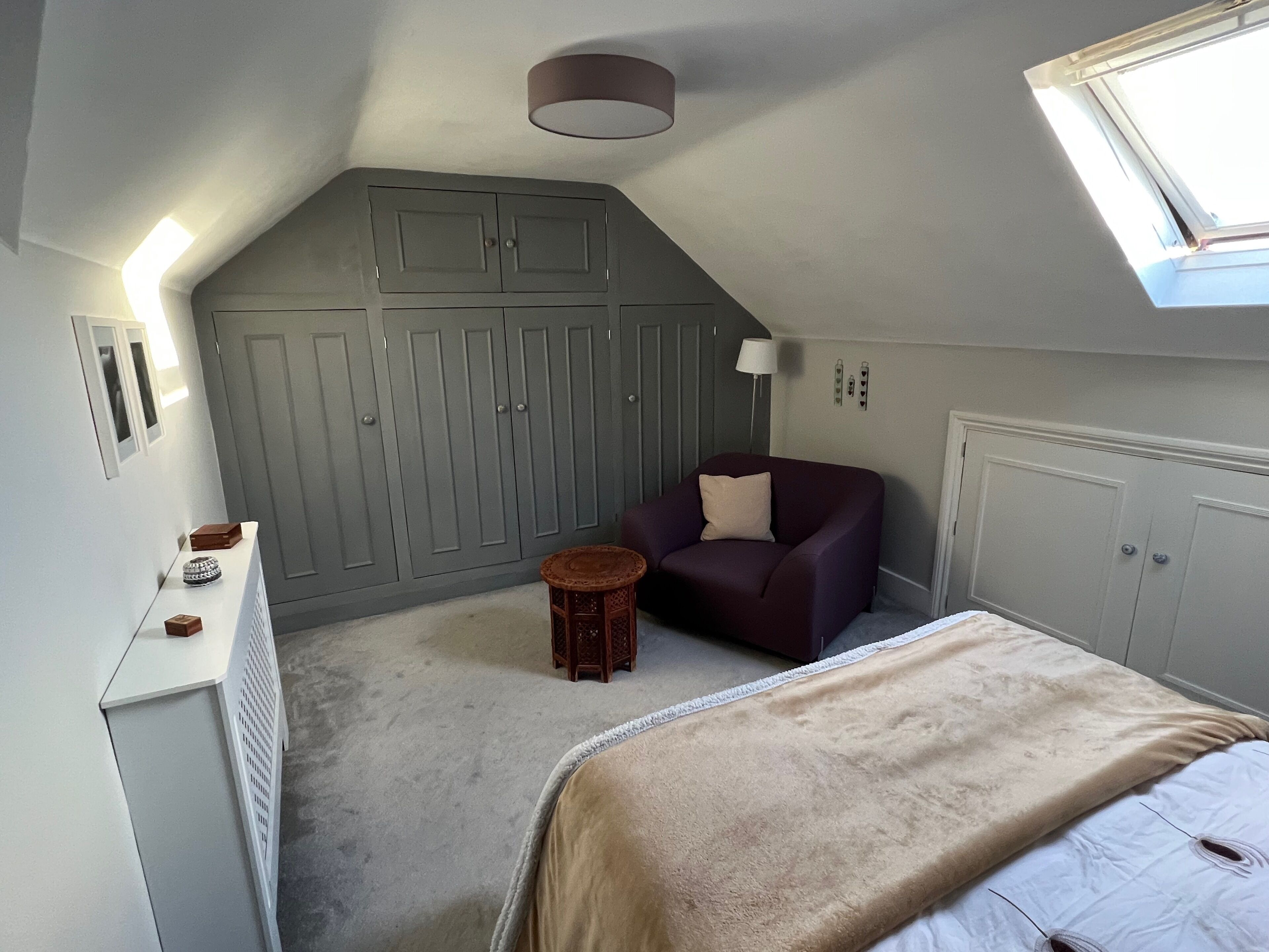 Une chambre avec un plafond en pente, un lit recouvert d’une couverture beige, un fauteuil violet, un petit tabouret en bois et une armoire grise.