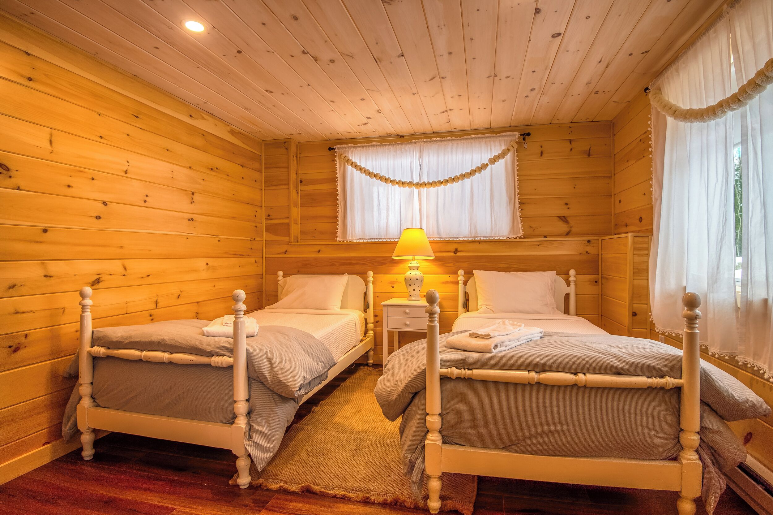 A cozy wooden cabin room with two single beds, a nightstand with a lamp, and a window with curtains.