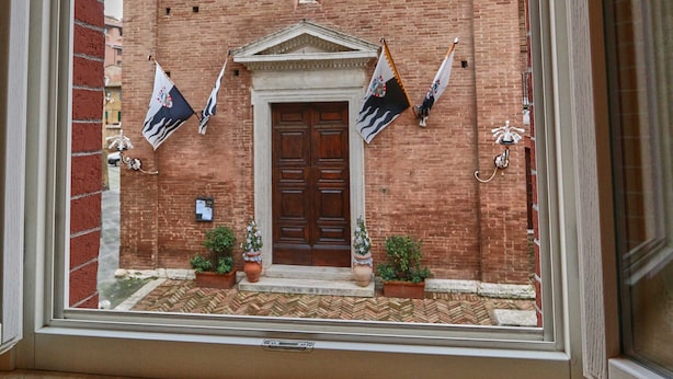 Vista attraverso una finestra di un edificio con una porta centrale, affiancata da due bandiere, e un finestrone circolare sopra la porta.