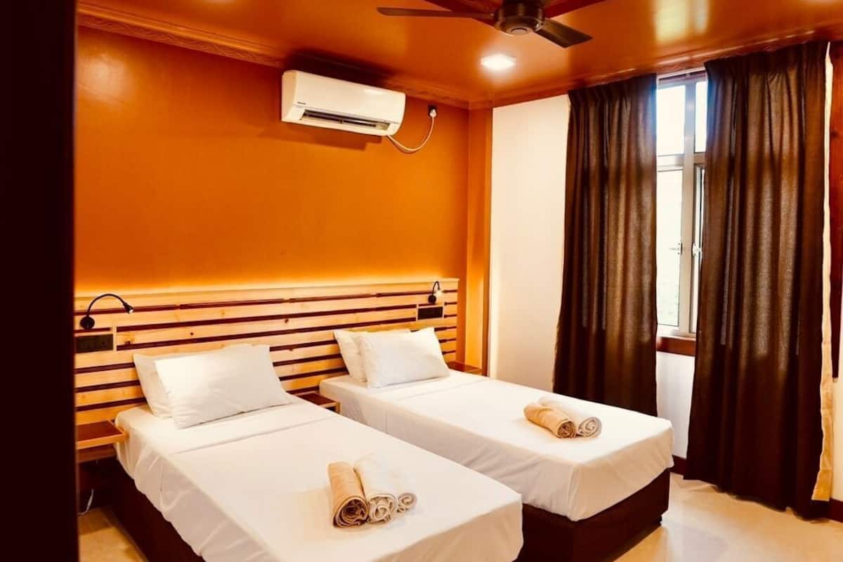 Habitación de hotel con dos camas, ventilador de techo, unidad de aire acondicionado y ventana con cortinas.
