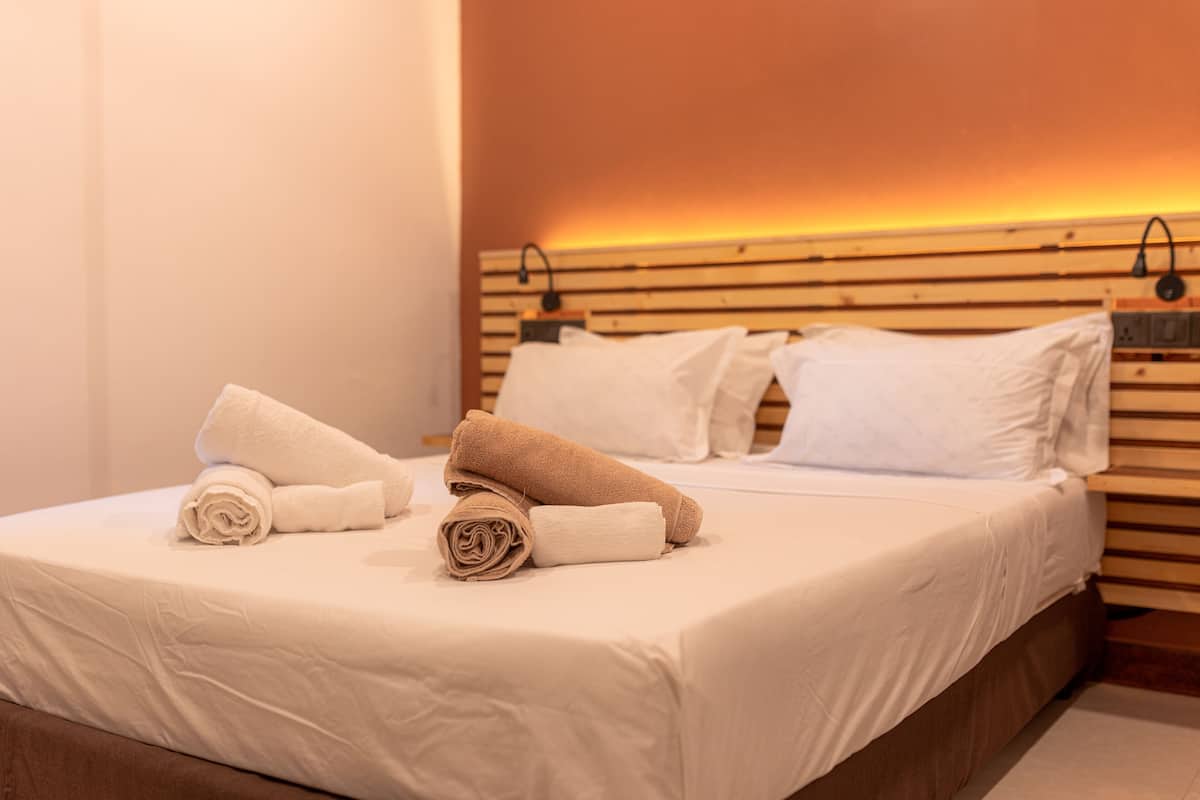 Una cama bien hecha con toallas blancas y beige, un cabecero de madera y una lámpara fijada a la pared.
