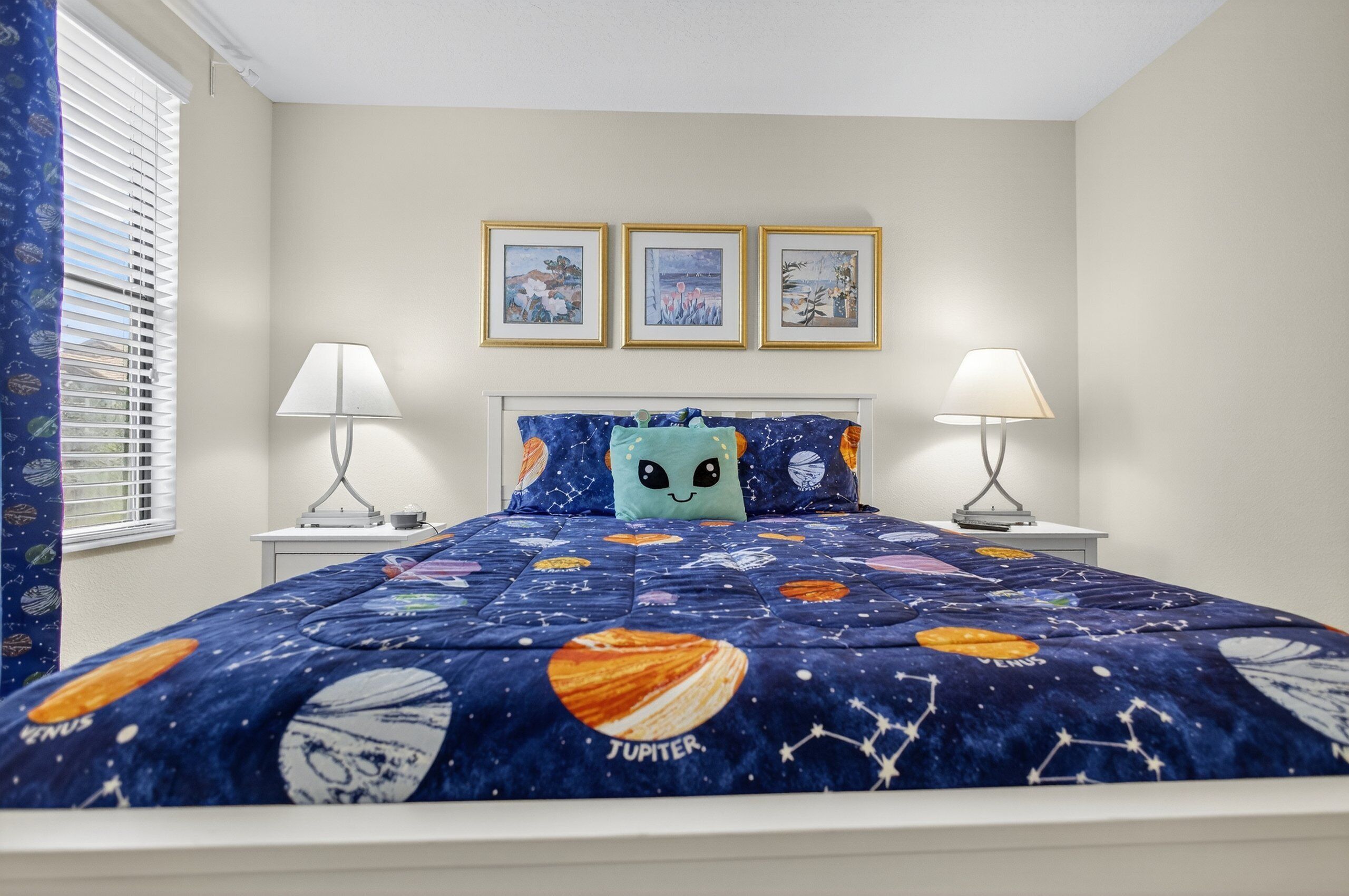 A bedroom with a bed covered in a space-themed quilt, two bedside lamps, and three framed pictures on the wall.