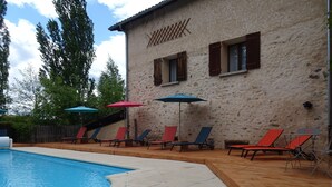 Una piscina con sillas de descanso y sombrillas, un edificio de piedra con contraventanas de madera, y una terraza de madera.