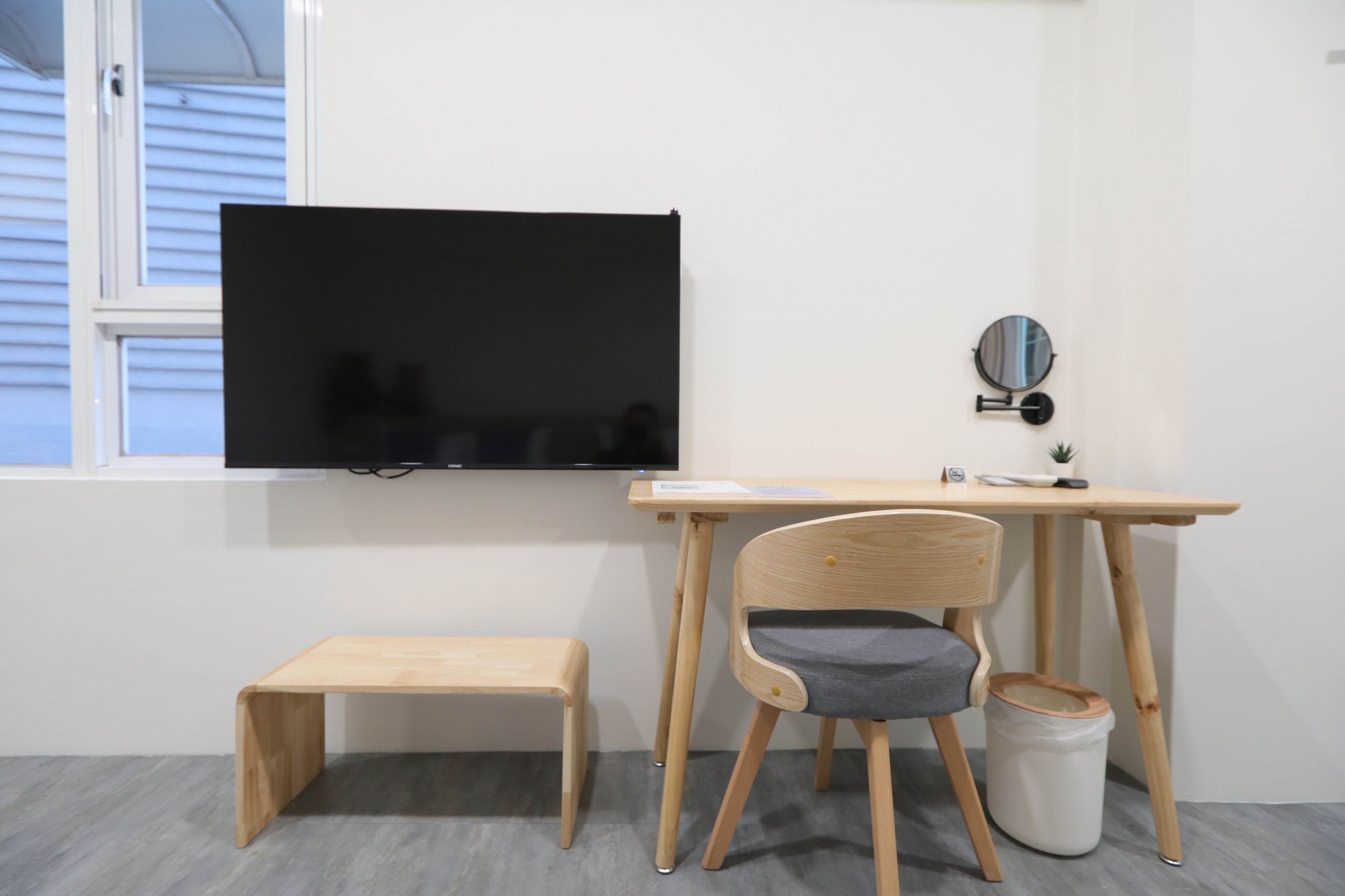 A room with a wooden desk, a chair, a small wooden bench, a television mounted on the wall, and a small round mirror.
