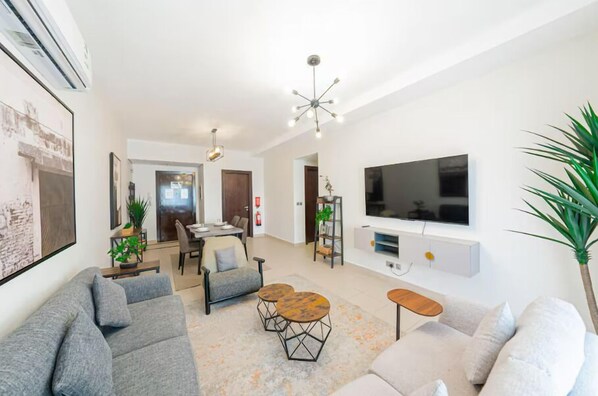 A modern living room with a grey sofa, a coffee table, a TV mounted on the wall, and a dining area with a table and chairs. - Mabaat - AL-Fayhaa - 514 (Jeddah)