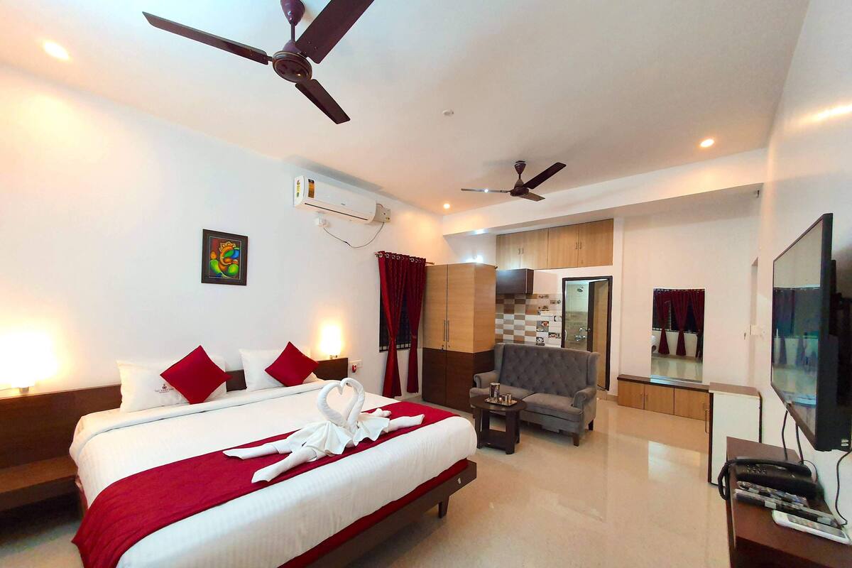 A hotel room with a large bed, two ceiling fans, a TV, a desk, and a bathroom visible through an open door.