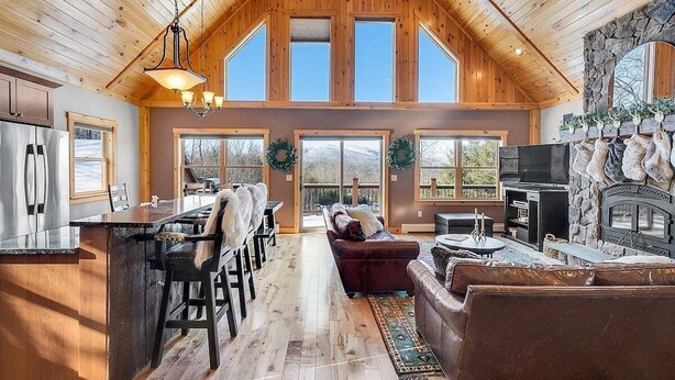 A spacious living area with a fireplace, a large window, and a stone wall. - Spruce Cliffs at Timber Creek (Bethel)