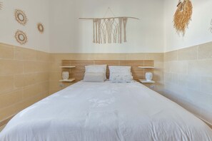 Cama bem arrumada com cabeceira de madeira, duas mesas de cabeceira com objetos decorativos e detalhes na parede, incluindo peças de macramé e arte circular.