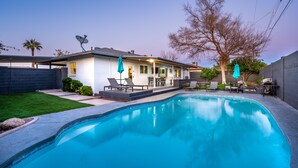 Una piscina con una casa al fondo, un patio con sillas y un árbol.