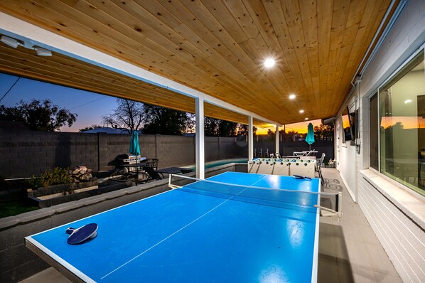 A ping pong table under a wooden ceiling with a pool table and foosball table nearby.
