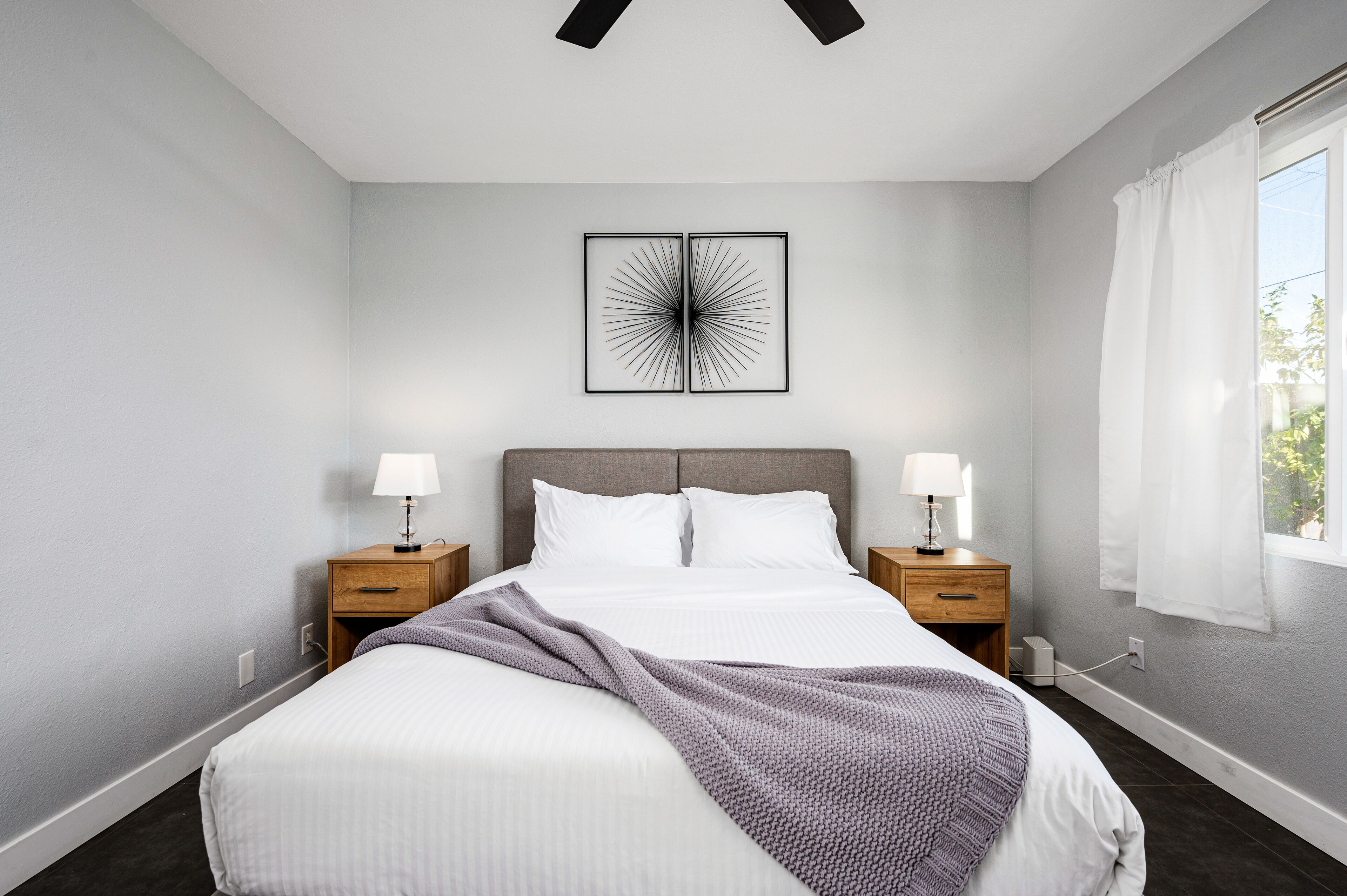 Quarto moderno com cama, duas mesas de cabeceira com luminárias, uma janela grande com cortinas e uma obra de arte abstrata na parede.