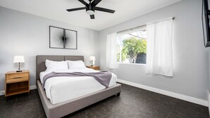 Un dormitorio moderno con cama, mesitas de noche, ventilador de techo, ventana con cortinas y televisor montado en la pared.