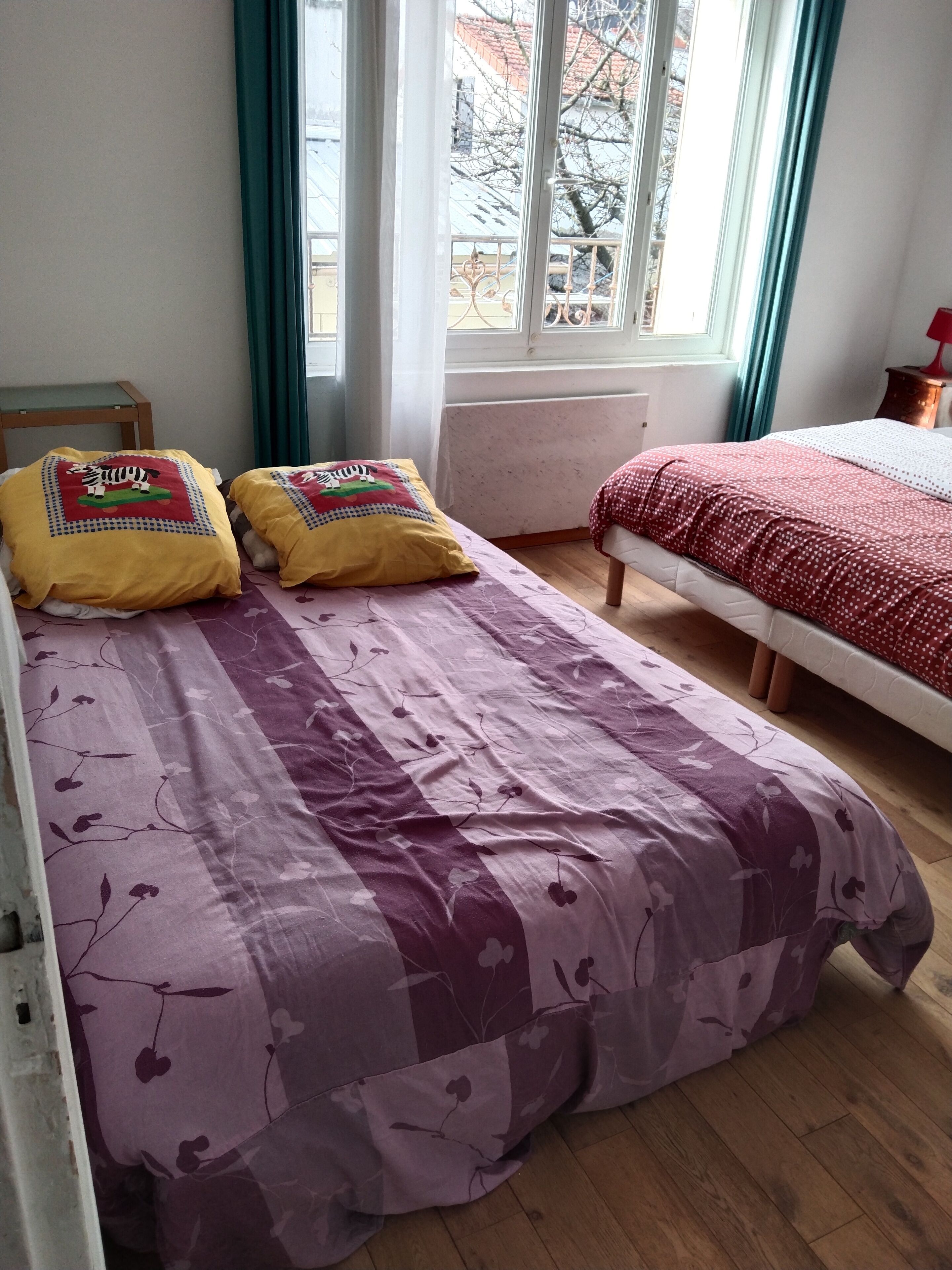 A bedroom with two beds, a window with curtains, a radiator, and a view of the outside.