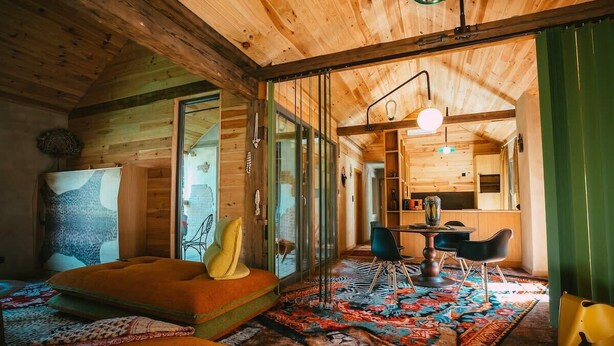 A cozy cabin interior with a wooden ceiling, a dining area with a round table and chairs, and a large window offering a view of the outdoors.
