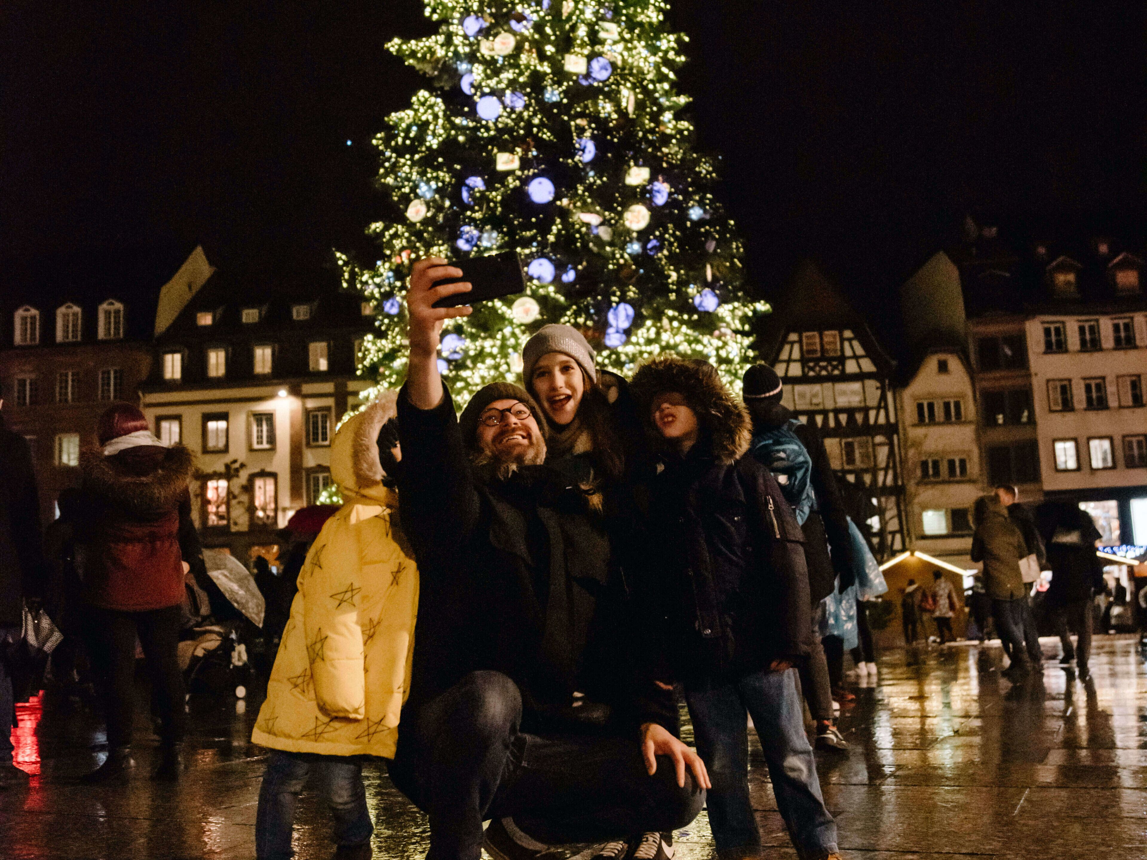 A group of people taking a selfie with a decorated Christmas tree in an urban square.