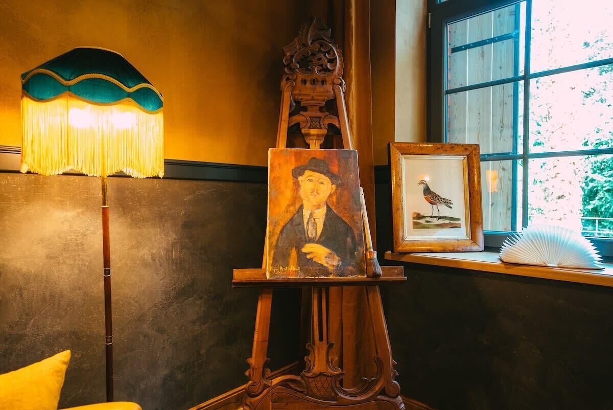 A wooden easel with a painting and a framed picture of a bird on a windowsill.