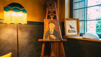 A wooden easel with a painting and a framed picture of a bird on a windowsill.