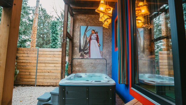 Ein Whirlpool in einem Holzdeckbereich mit einem Bild einer Frau im Kleid, einem Holzzaun und einer Glastür, die Bäume reflektiert.