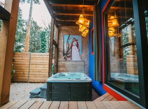 A hot tub in a wooden deck area with a painting of a woman in a dress, wooden fence, and a glass door reflecting trees. - Viva Frida ! Unique accommodation with private jacuzzi in Alsace (Guebwiller)
