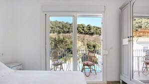 Ein Schlafzimmer mit Bett, Nachttisch, einem Balkon mit Korbstühlen und Blick auf Pool und Bäume.
