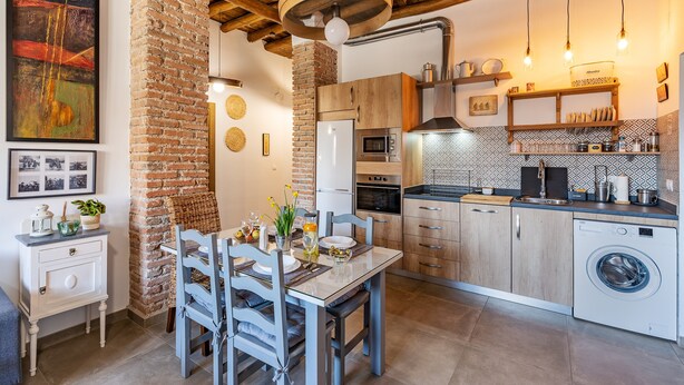 A modern kitchen with a brick wall, wooden ceiling beams, and a dining area with a table and chairs. - Country House 'La Criba - Casa Rural Eralta' with Mountain View, Wi-Fi and Air Conditioning (Dúrcal)