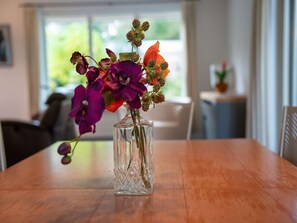A clear glass vase with purple and orange flowers on a wooden table in a room with a window and a plant in the background. - Free Holiday Home With big Garden (Schoonloo)