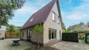 Een huis met twee verdiepingen, een schuin dak, een kleine tuin met een tafel en een jonge boom, en een haag aan de zijkant.