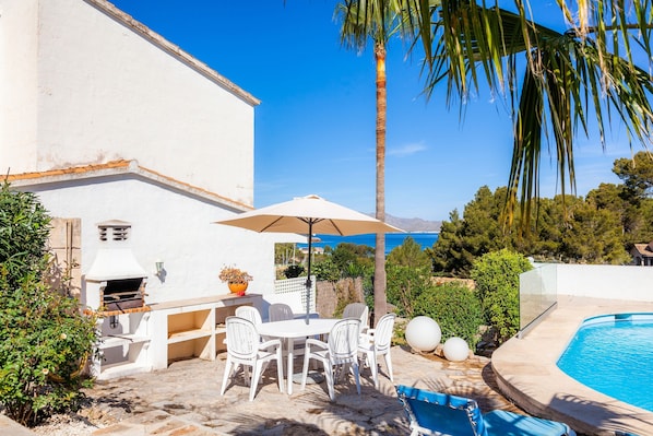 Une terrasse avec un barbecue, des chaises blanches et une table placée sous un parasol, d’où l’on peut admirer une piscine et un ciel dégagé.