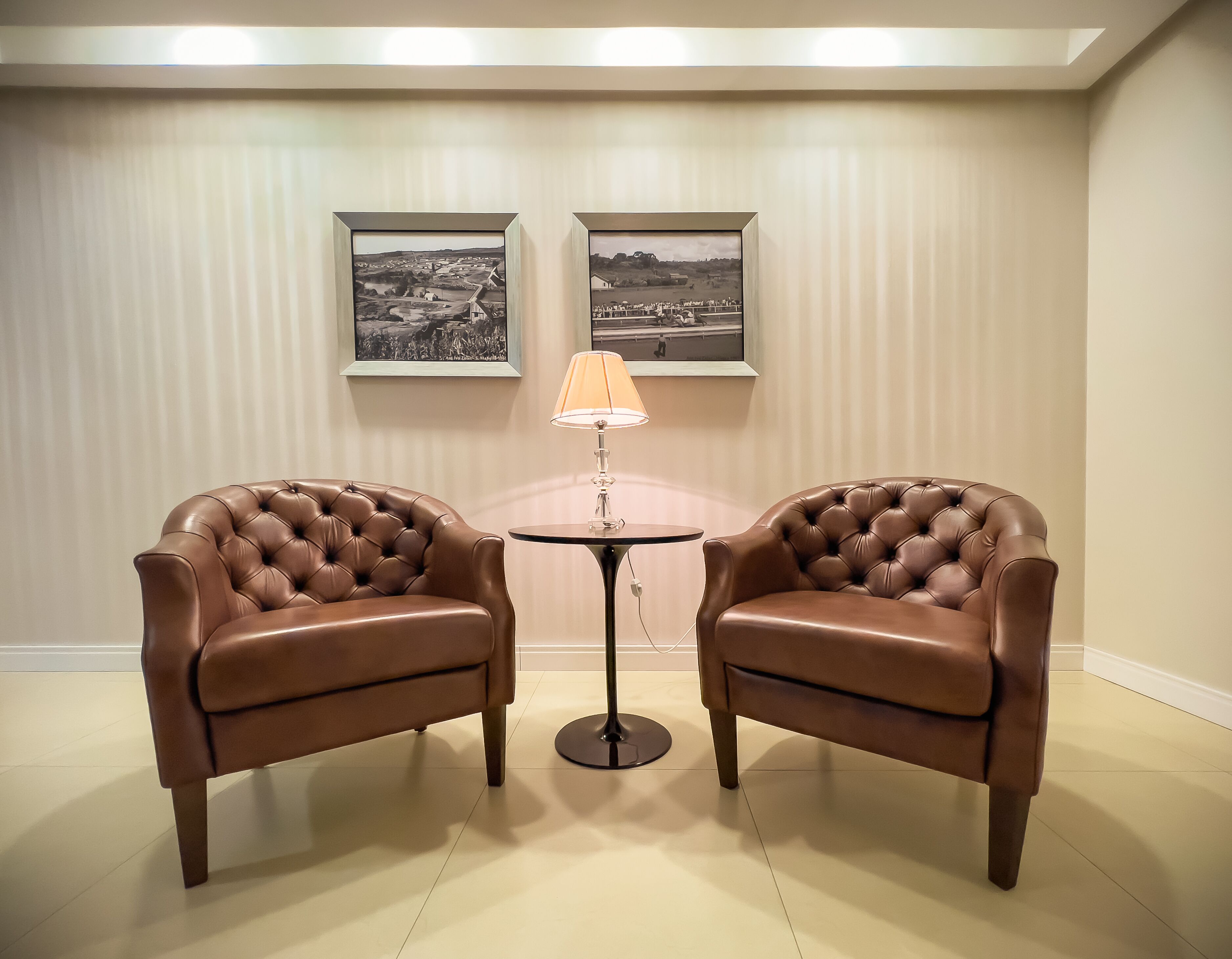 A room with two brown leather armchairs, a small round table with a lamp, and two framed black-and-white photographs on the wall.