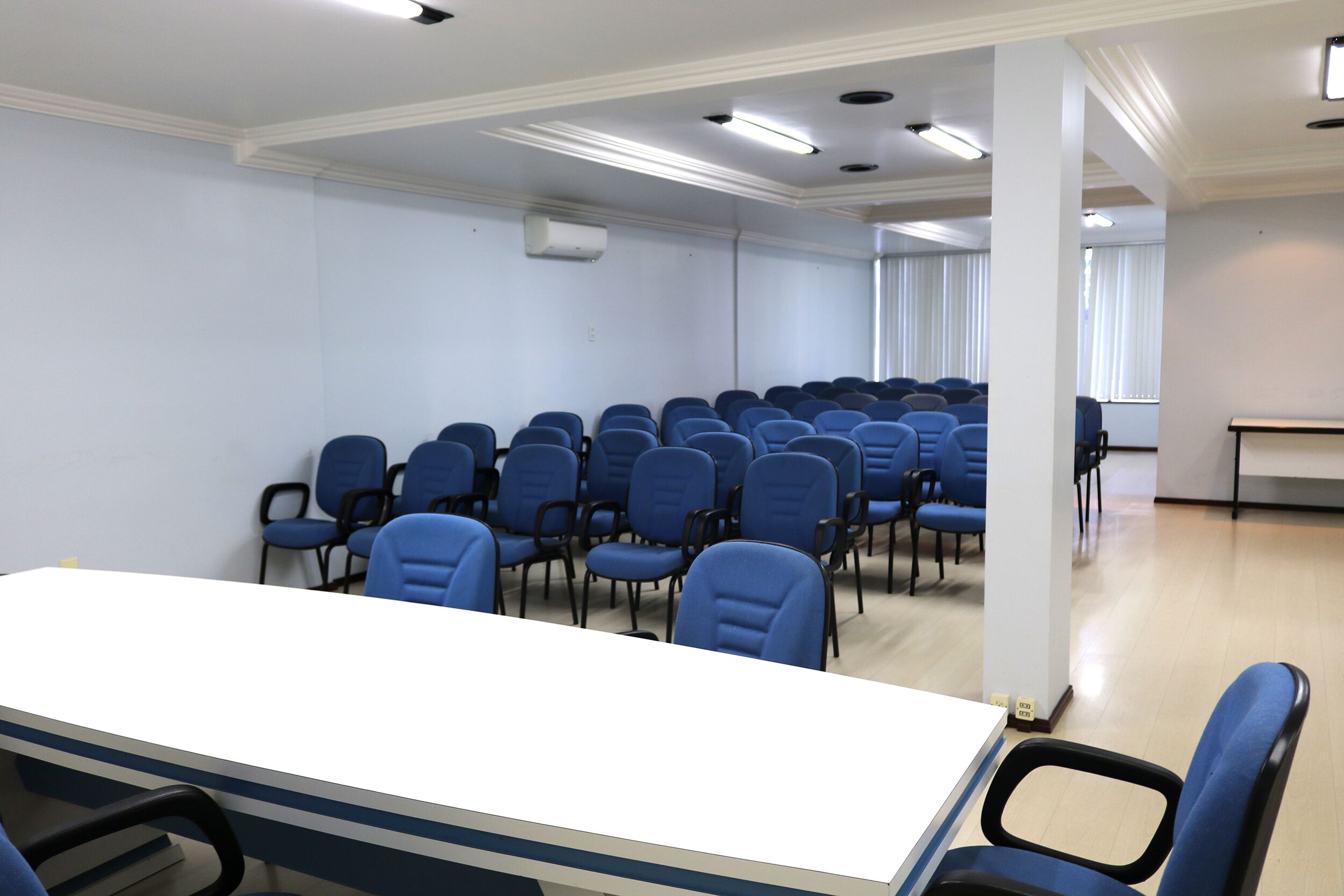 A conference room with rows of blue chairs, a white table, and a large window with blinds.