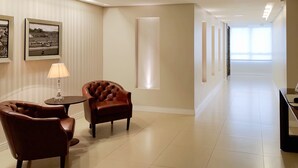 A hotel corridor with a leather armchair and a small table with a lamp, framed pictures on the wall, and a long, well-lit hallway. - Tri Hotel Xanxerê (Xanxere)