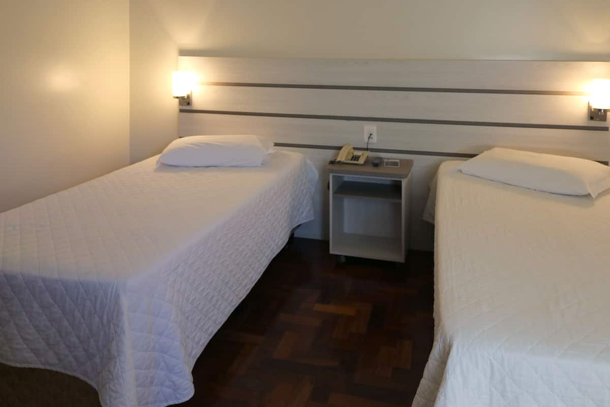 Ein Hotelzimmer mit zwei Einzelbetten, jeweils mit weiß gestopfter Bettdecke und Kissen. An den Nachttischen stehen jeweils eine Lampe und ein Telefon. Der Boden ist mit Holzparkett belegt und das Kopfteil weist horizontale Streifen auf.