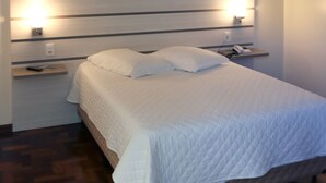 A neatly made bed with white bedding, a bedside table with a lamp, and a wall-mounted light fixture. - Tri Hotel Xanxerê (Xanxere)