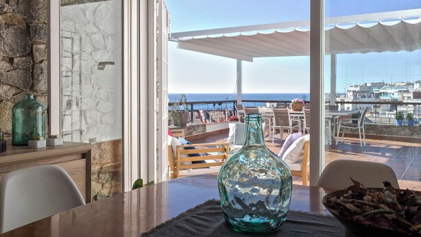 Una terraza con muro de piedra, una mesa de madera, una botella de vidrio y vistas al mar y edificios.