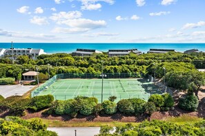 A tennis court surrounded by lush greenery and buildings with a view of the ocean.