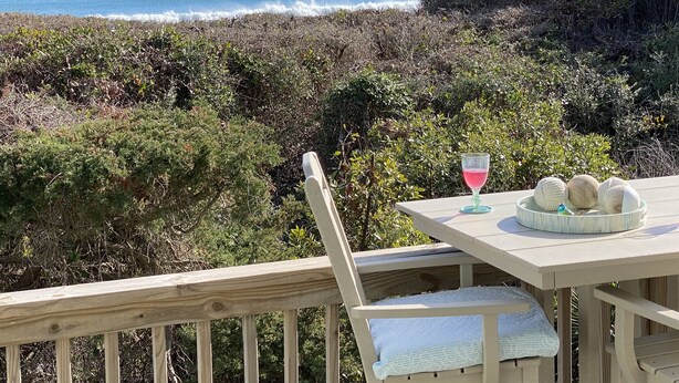 Een houten terras met een tafel en stoel met uitzicht op een strand met golven en weelderige begroeiing.