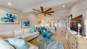 A spacious living room with a sofa, teal armchair, and a colorful rug. A ceiling fan and kitchen are visible in the background.