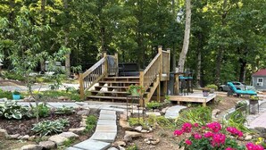 A wooden deck with a staircase leading to a seating area, surrounded by a garden with various plants and flowers.